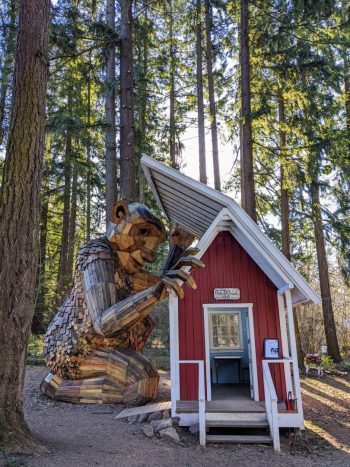 A large wooden bald troll made from deconstructed shipping palettes holds open one side of a small red shed's pointy roof to look inside of it. (The shed was clearly modified to allow the troll statue to do this.) The troll is surrounded by tall evergreen trees.