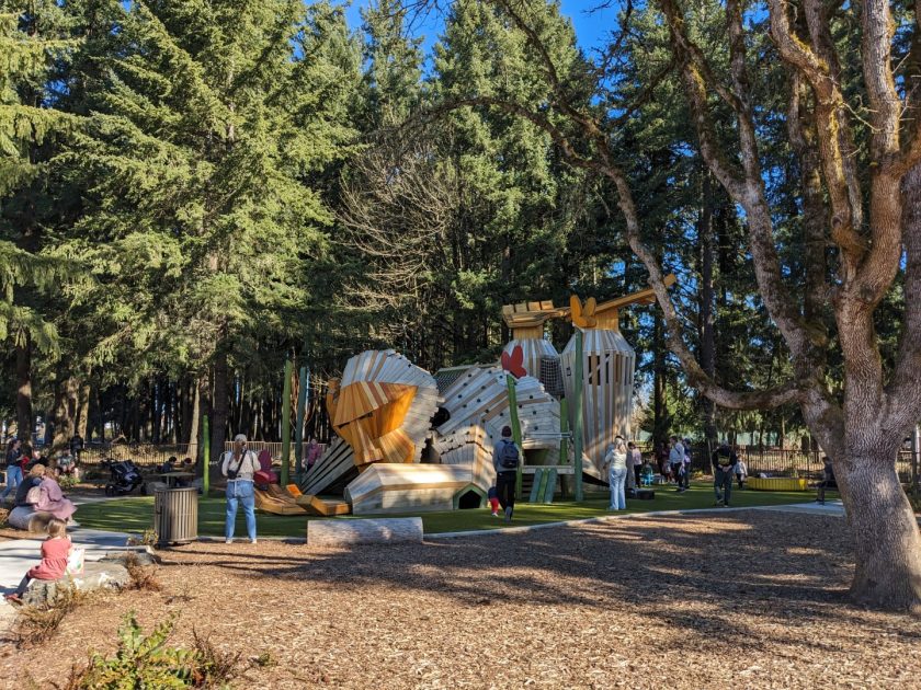 A large play structure, approximately 15 feet tall and 40 feet long that is shaped like a monkey lying on its stomach, looking at a butterfly on it's finger, with it's legs bent at the knees and feet high in the air. Children and adults surround, climb on, and enter the structure.