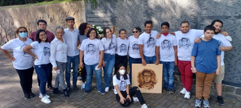 Sixteen people pose in front of a memorial wall wearing white shirts with an illustration of Efraín’s face.